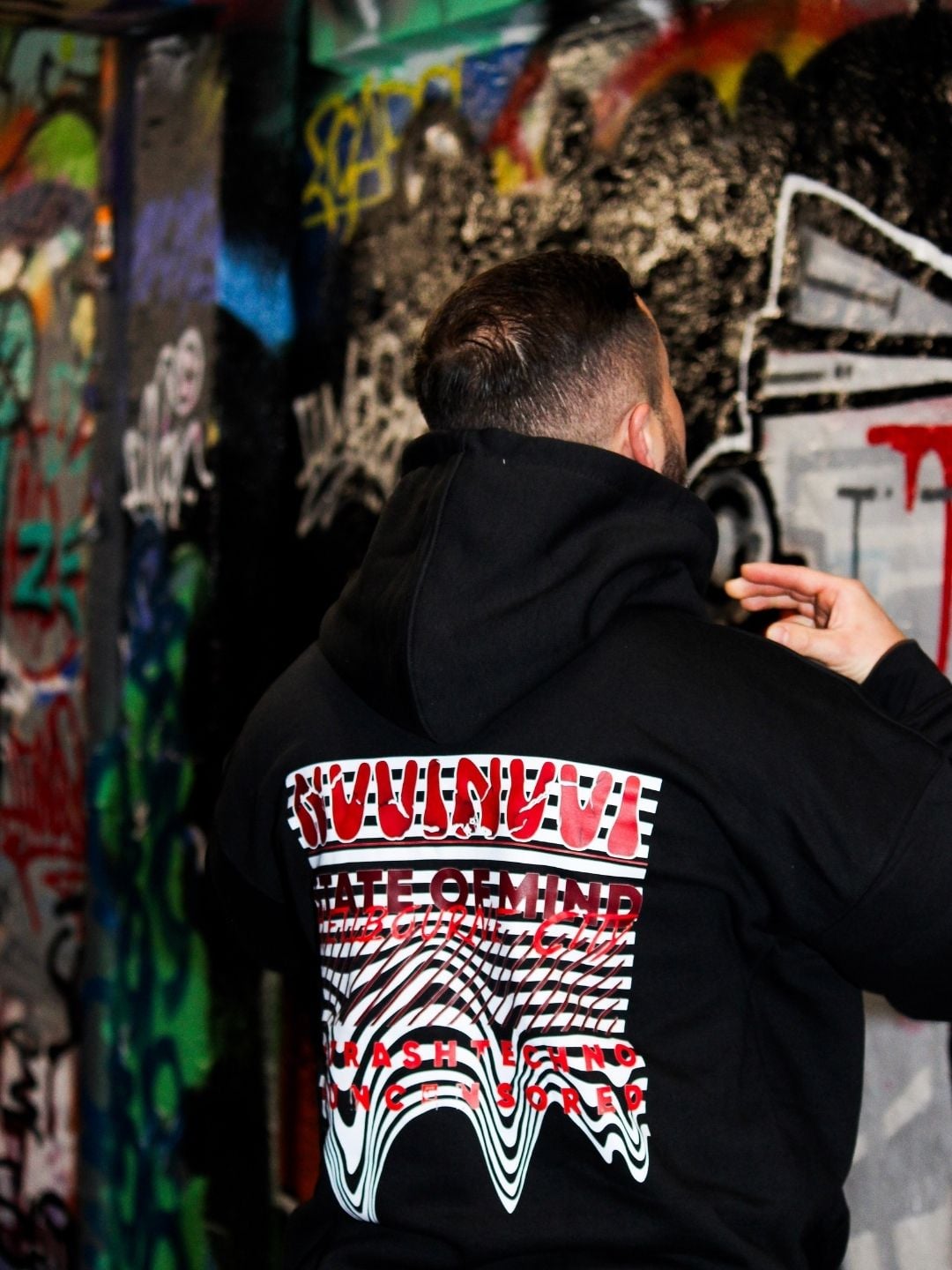 Black hoodie worn from behind, melted red and white NVVINVVI back print, against a graffiti wall.