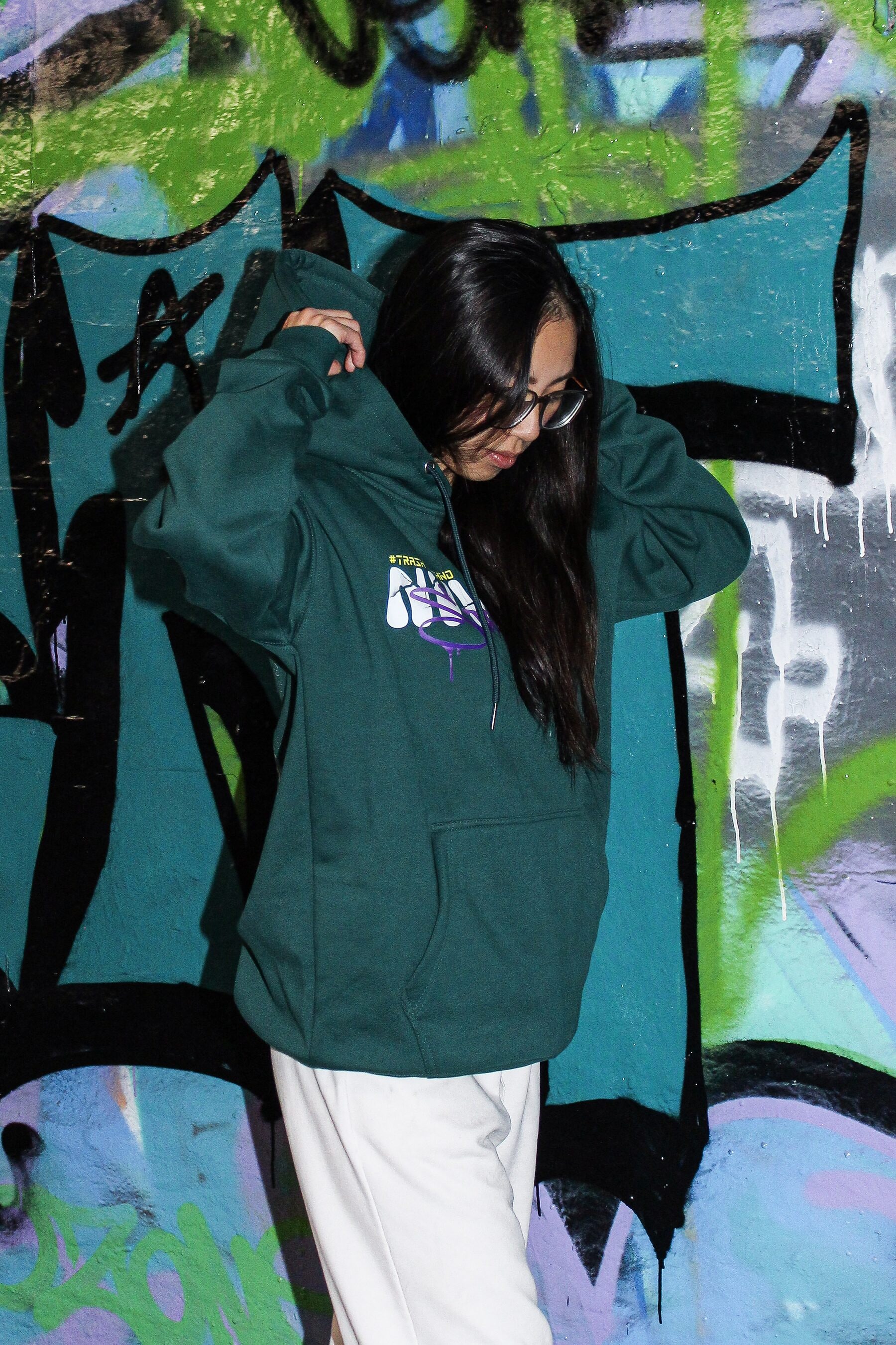 VINV forest-green hoodie worn from the front, hood pulled up with both hands, against a turquoise graffiti wall