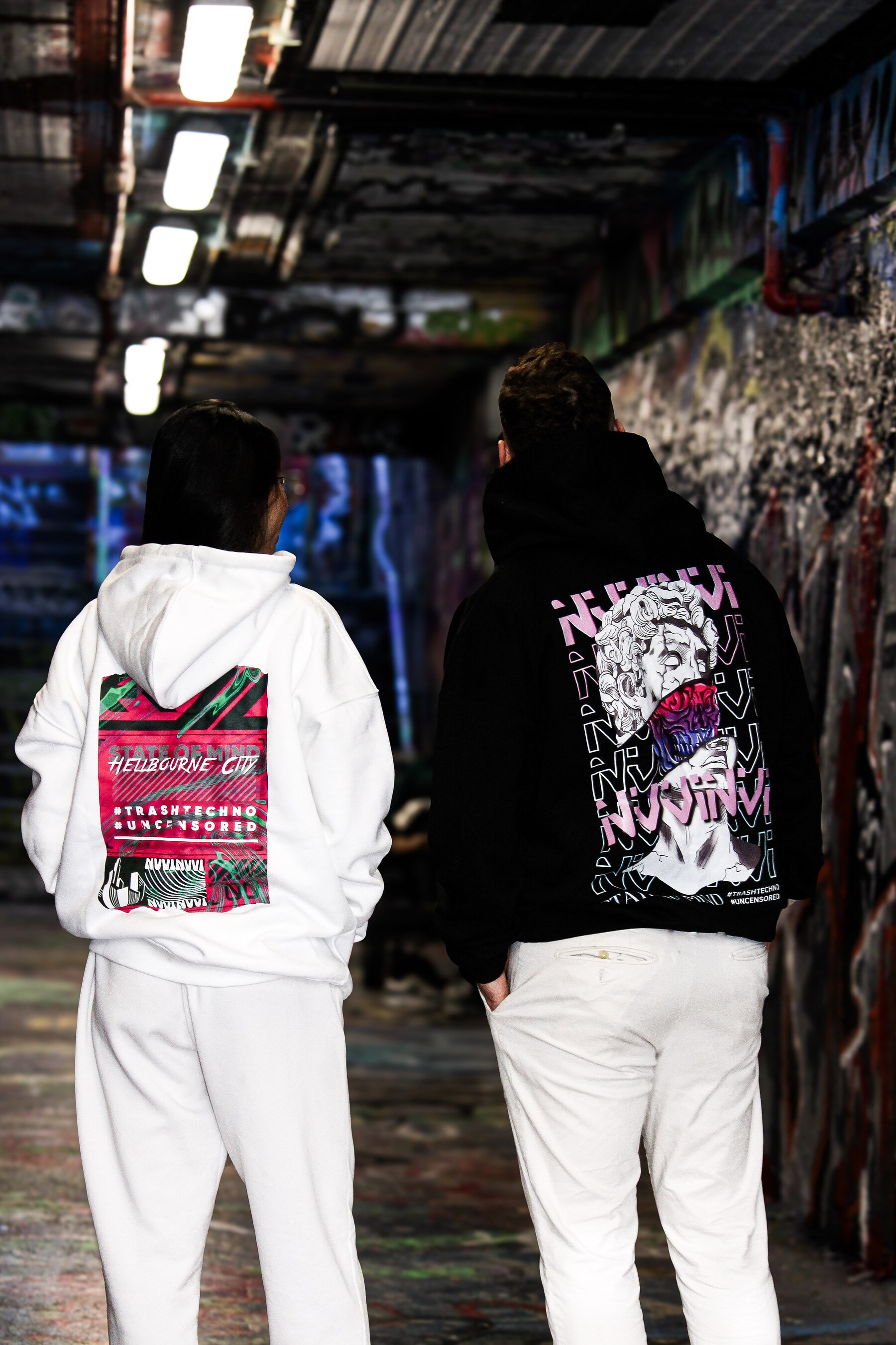 Two VINV hoodies worn from the back, white and black back prints, in a graffiti-covered tunnel
