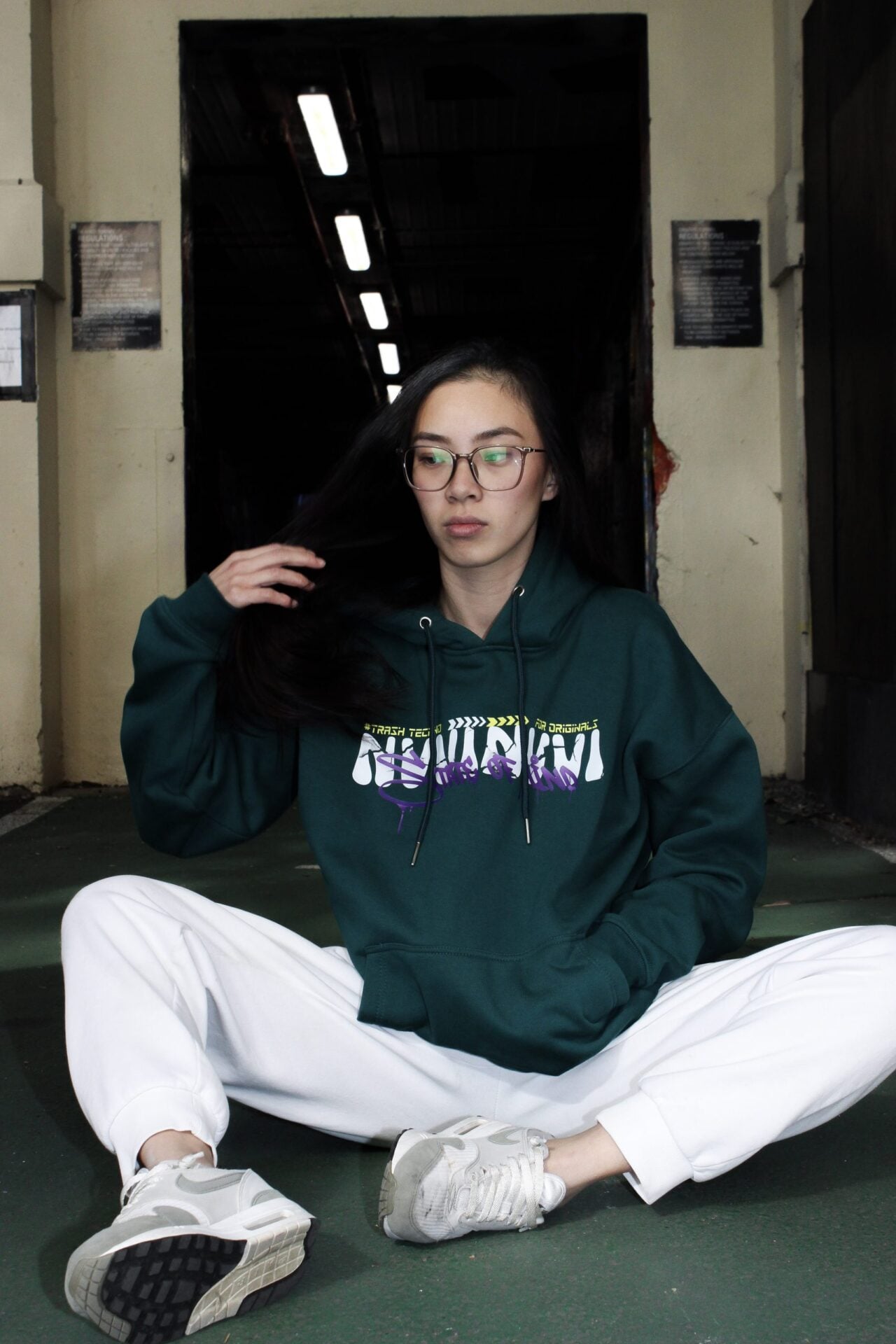 Hoodie vert porté de face, lettrage NVVINVVI sur la poitrine, assise au sol.