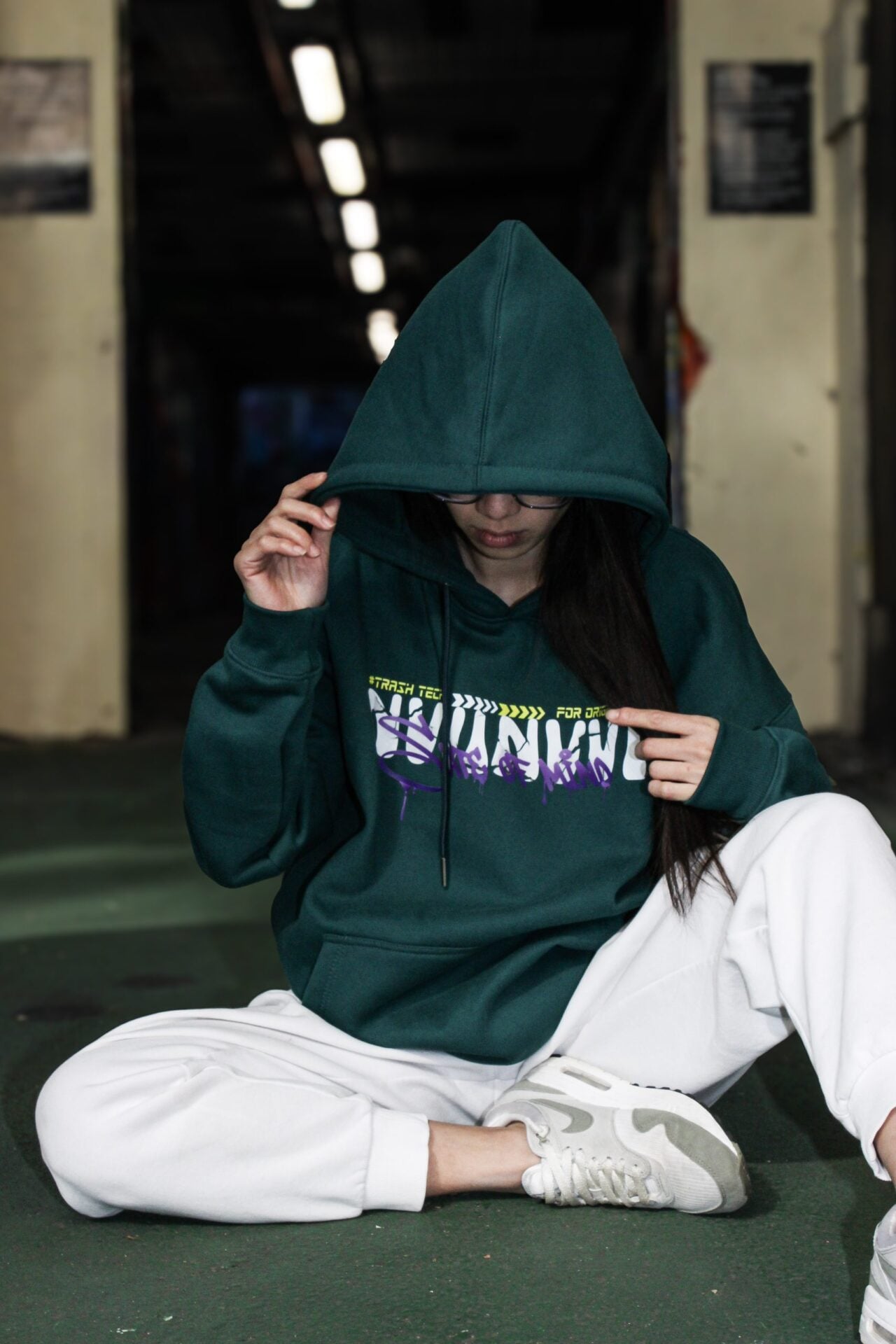 Hoodie vert porté capuche relevée, assise au sol dans un hall sombre.