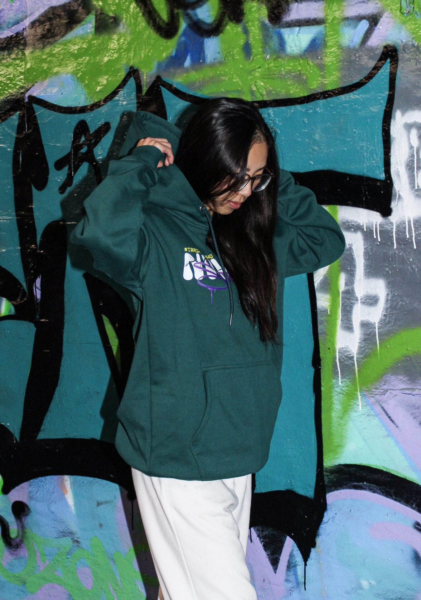 Hoodie vert porté de face devant un mur de graffitis.