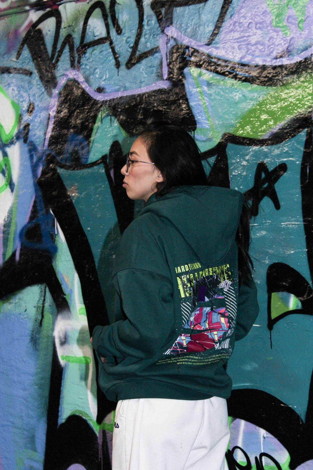 Hoodie vert vu de trois quarts dos devant un mur de graffitis, impression dorsale visible.