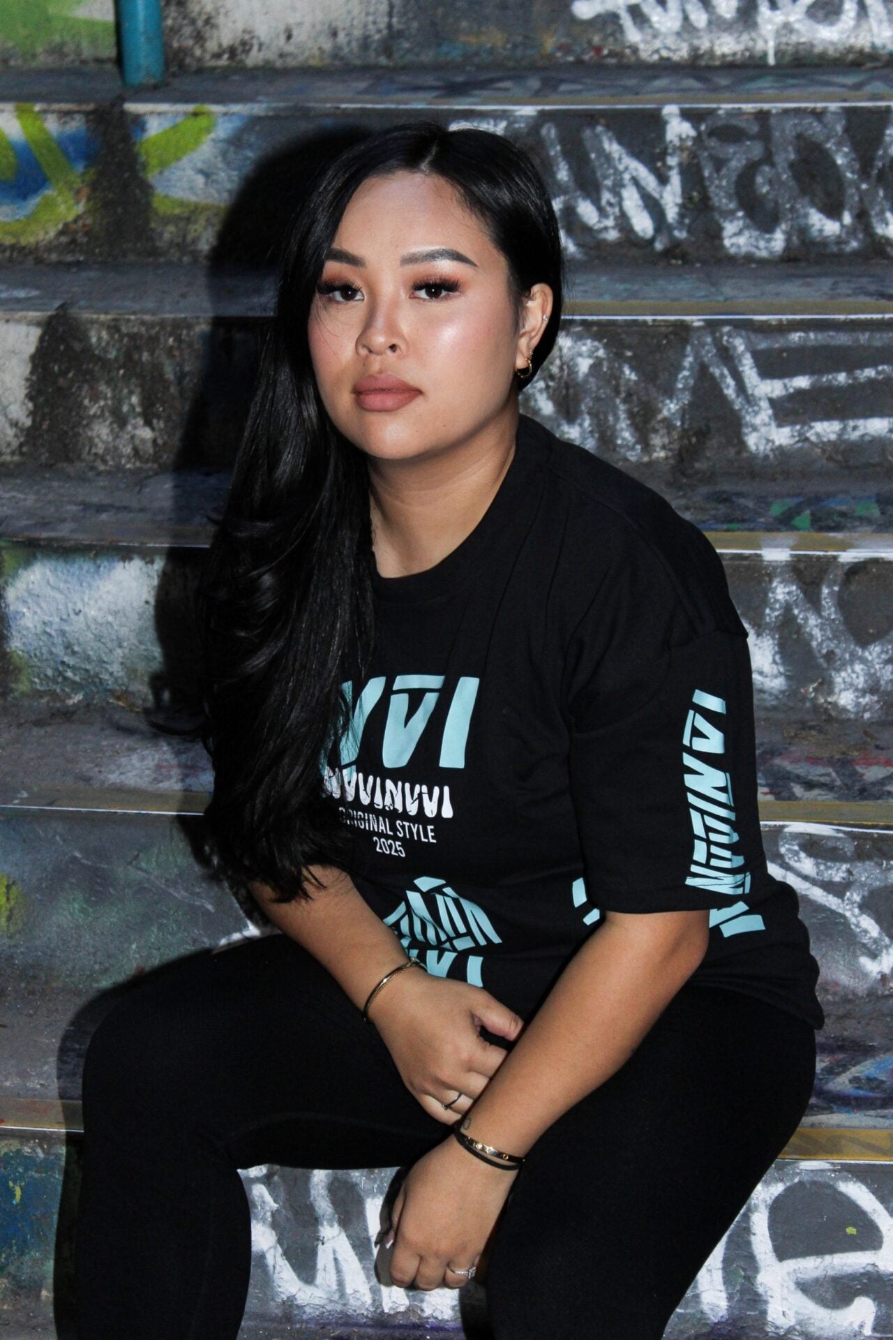 T-shirt noir porté de face, lettrage cyan, assise sur un escalier.