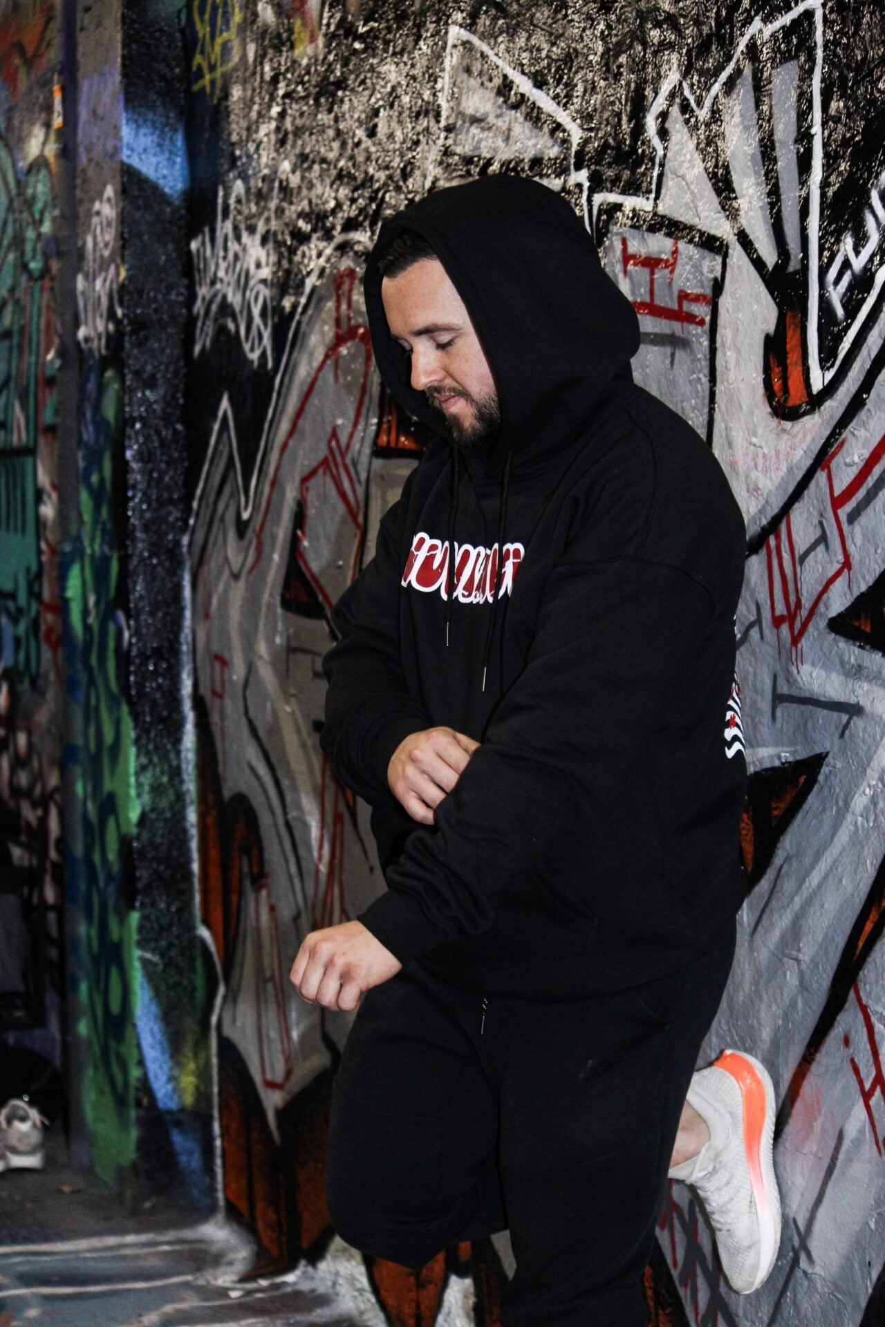 Hoodie noir capuche relevée, vue de trois quarts, dans un passage couvert de graffitis.