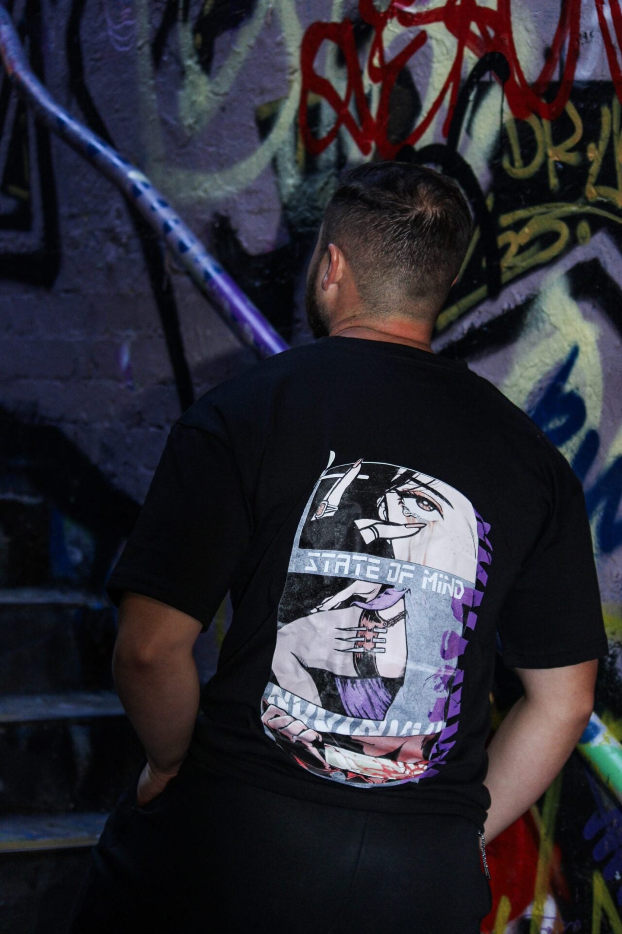Dos du t-shirt noir, impression State of Mind violette, devant un mur de graffitis.