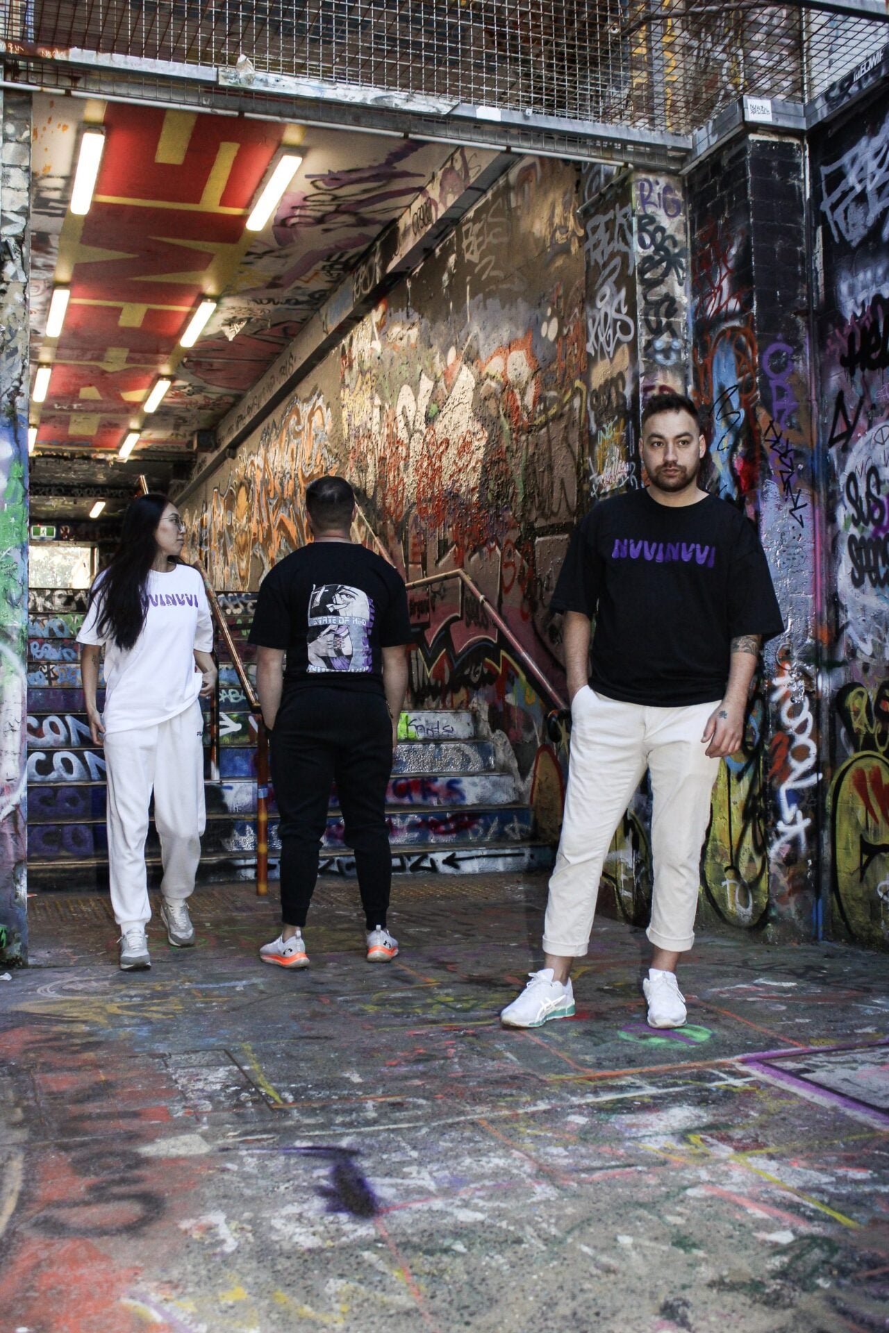 White and black versions of the tee, seen from behind and from the front, in a graffiti-covered tunnel.