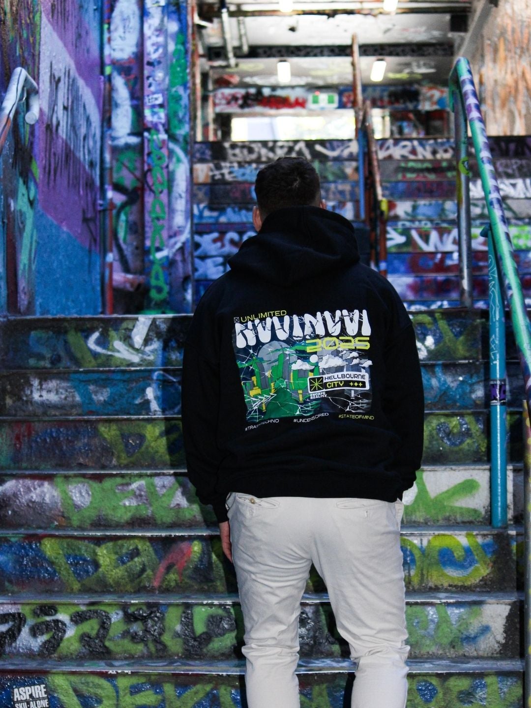Hoodie noir porté de dos sur un escalier couvert de graffitis, impression dorsale verte et blanche.